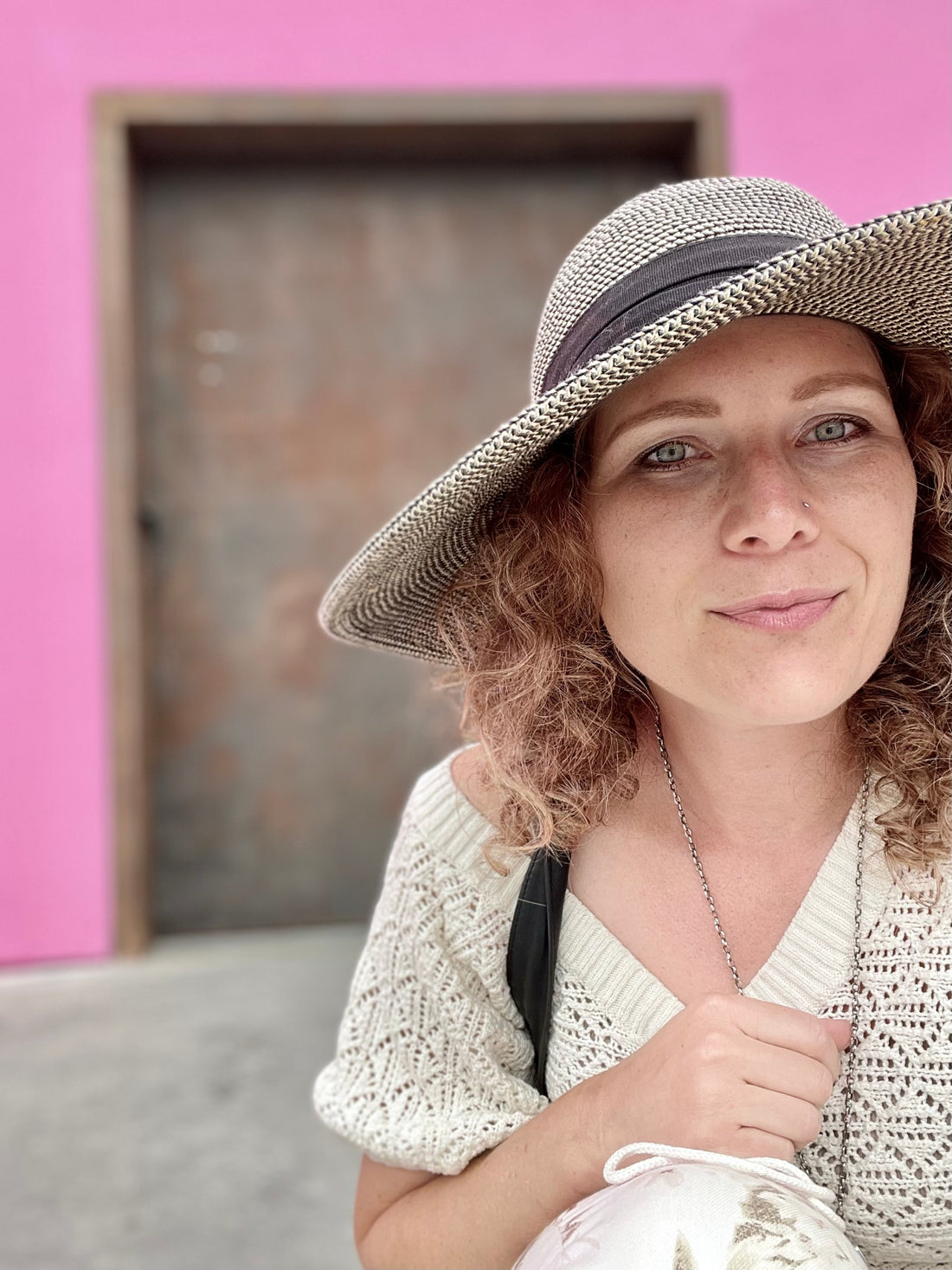 The image features a close-up of Heather outdoors with a bright pink wall and a blurred door in the background. Light complected, Heather has light curly hair and is wearing a wide-brimmed, textured straw hat with a dark band. She is dressed in a cream-colored, knit top with a delicate, lacy pattern. Heather has a relaxed, gentle expression with a subtle smile and a small nose piercing. The background contrasts sharply with the person's light-colored clothing and hat, creating a striking visual effect. 